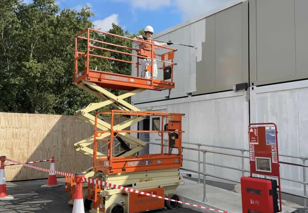 Construction worker in a hard hat on an orange elevated scissor lift applying paint to a gray building wall outside.